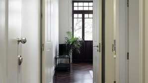 Un couloir aux portes blanches et au plancher en bois, qui mène à une pièce dotée d’une grande fenêtre et d’une plante.