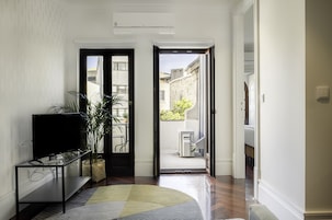 A hallway with a television on a glass table, a potted plant, and a rug leading to an open door to a balcony with a view of buildings and a tree.