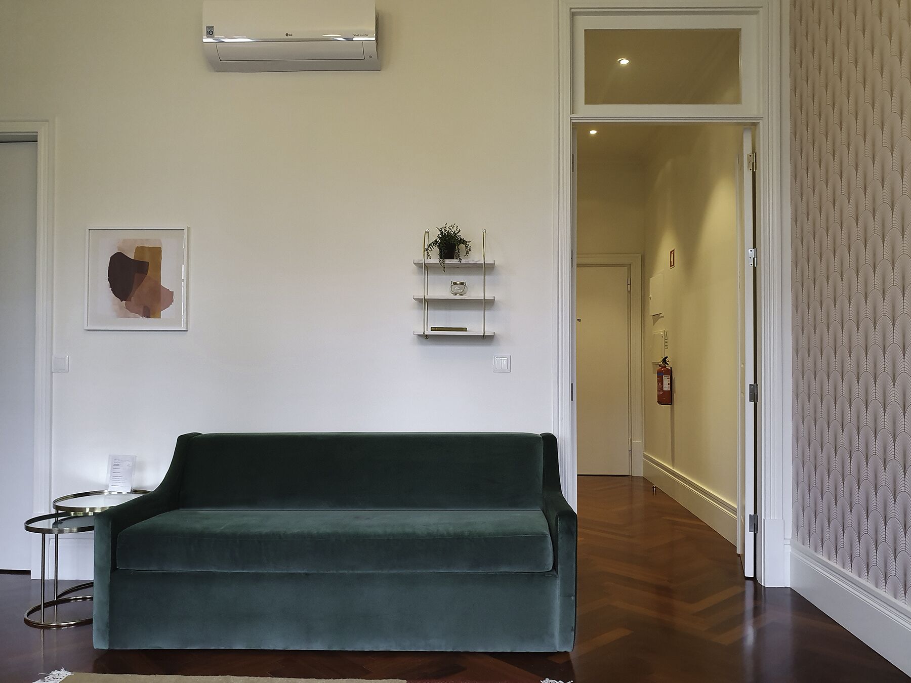 Un salon avec un canapé vert, une petite table d’appoint, une œuvre d’art abstraite encadrée, une étagère murale ornée d’objets décoratifs et un papier peint à motifs sur le mur de droite.