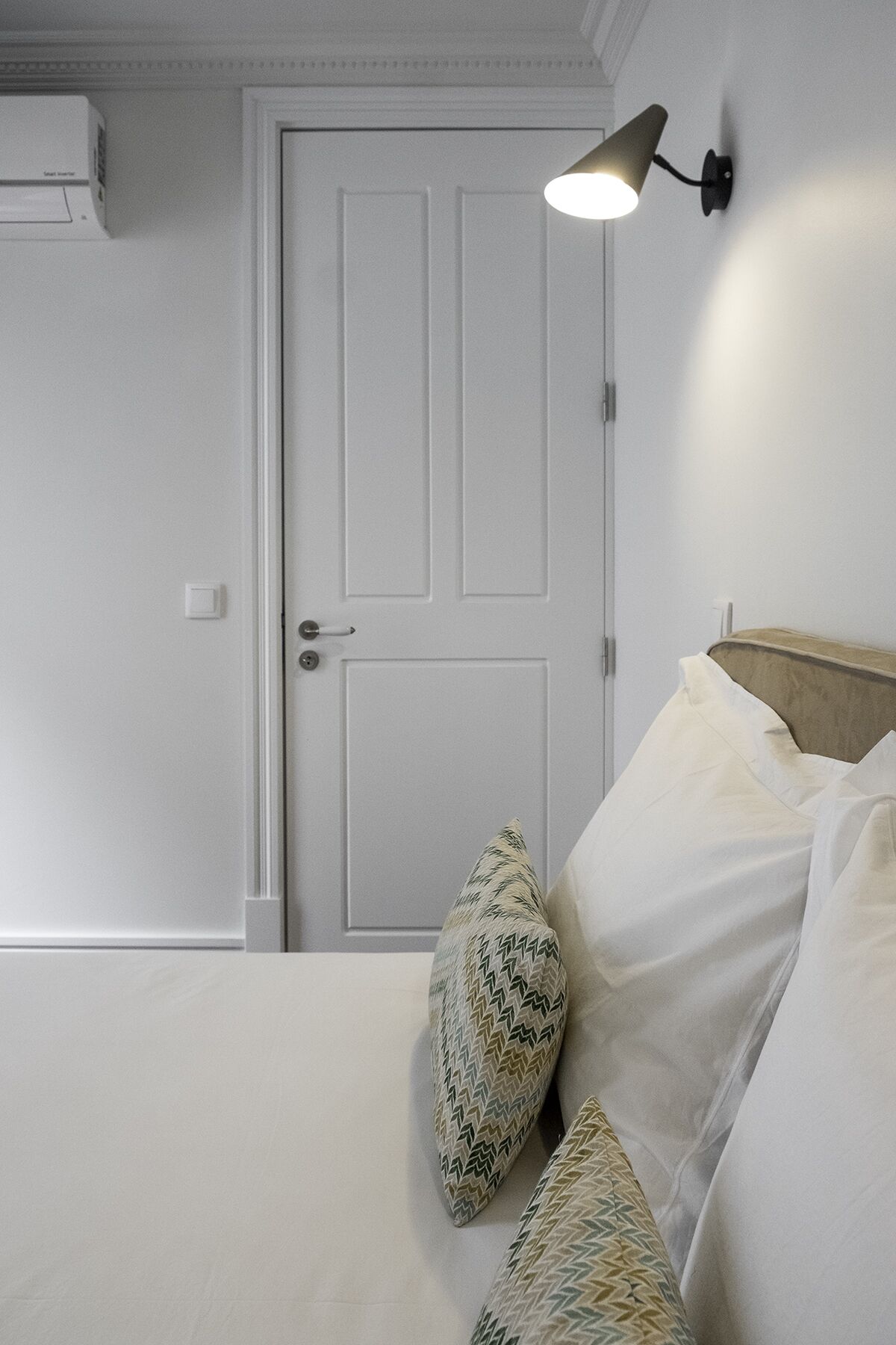Un dormitorio con una puerta blanca, una cama con ropa blanca y una lámpara fijada en la pared.