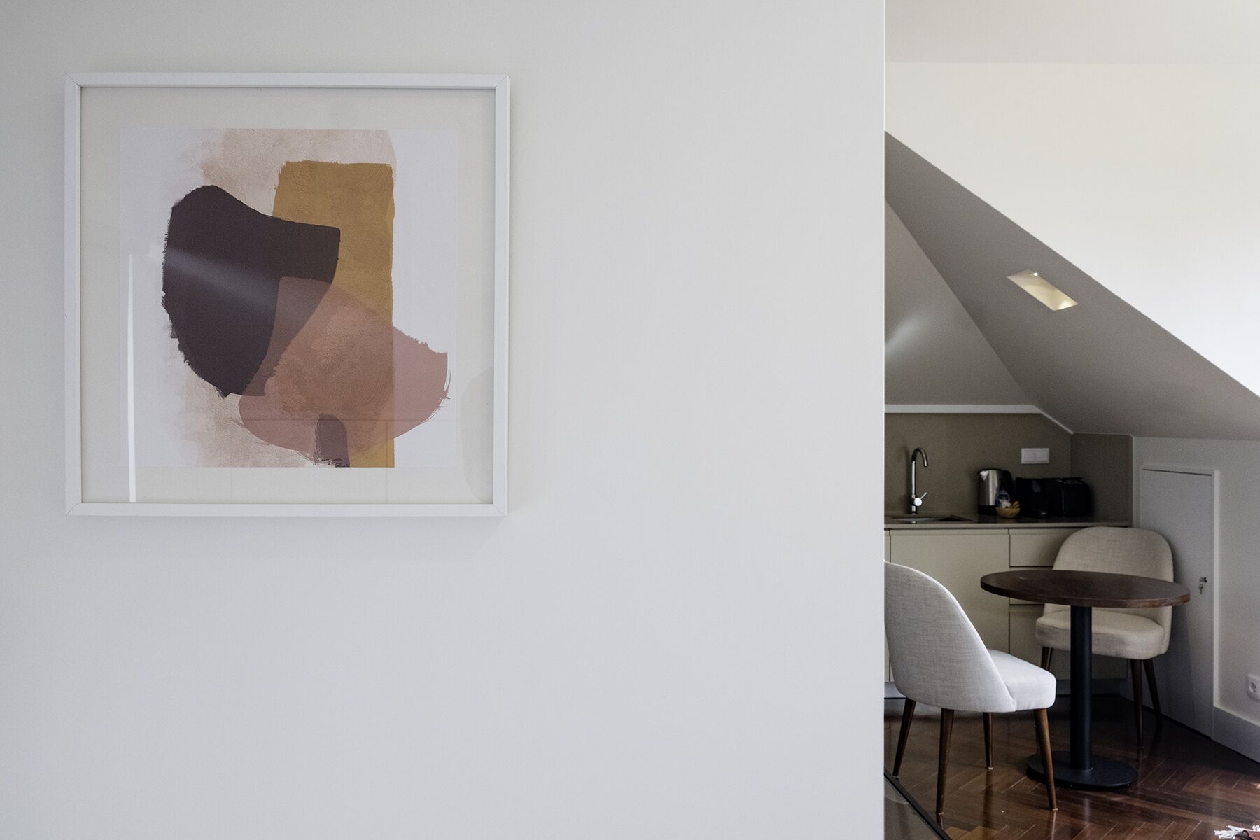 A modern interior with a framed abstract painting, a dining area with a round table and chairs, and a kitchen with a sink and countertop appliances.