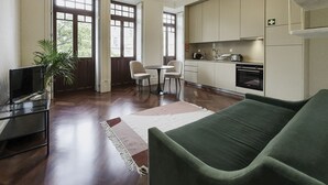Un salon moderne avec un canapé vert, une table à manger avec des chaises, un téléviseur posé sur un meuble et un coin cuisine équipé de placards et d’appareils électroménagers.