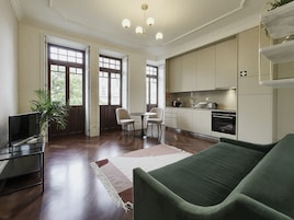 A modern living room with a green sofa, a dining table with chairs, a TV on a stand, and a kitchen area with cabinets and appliances.