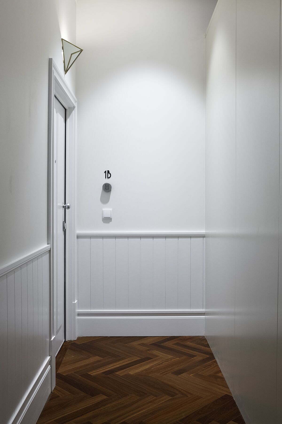 Un couloir aux murs blancs, doté d’un plancher en bois, d’une porte et d’un luminaire fixé au mur.