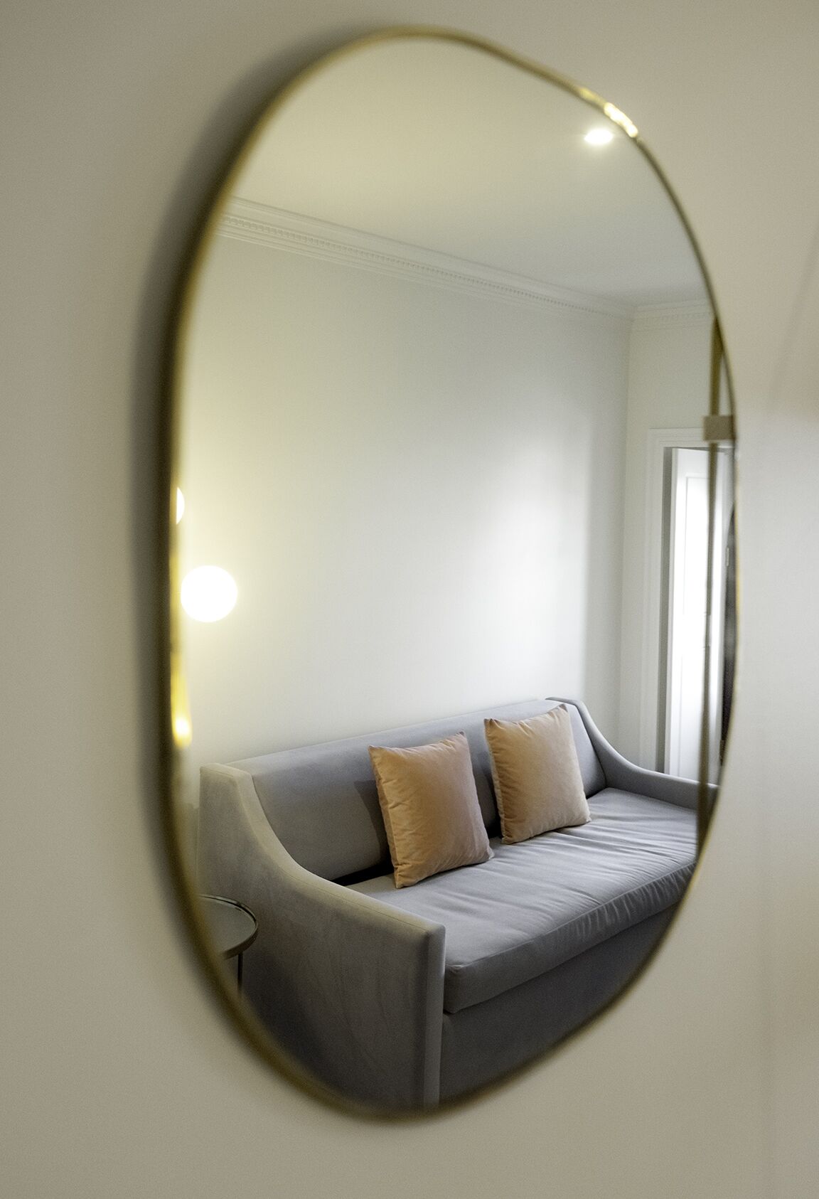 A modern room with a grey sofa, two beige cushions, and a large oval mirror on the wall.