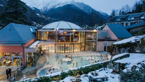 Um resort de spa com piscina central, áreas envidraçadas e montanhas ao fundo.