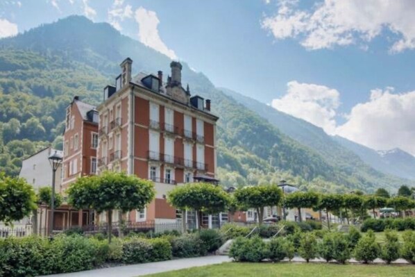 Un edificio grande de ladrillo rojo con múltiples ventanas y un balcón, rodeado de abundante vegetación y montañas al fondo.