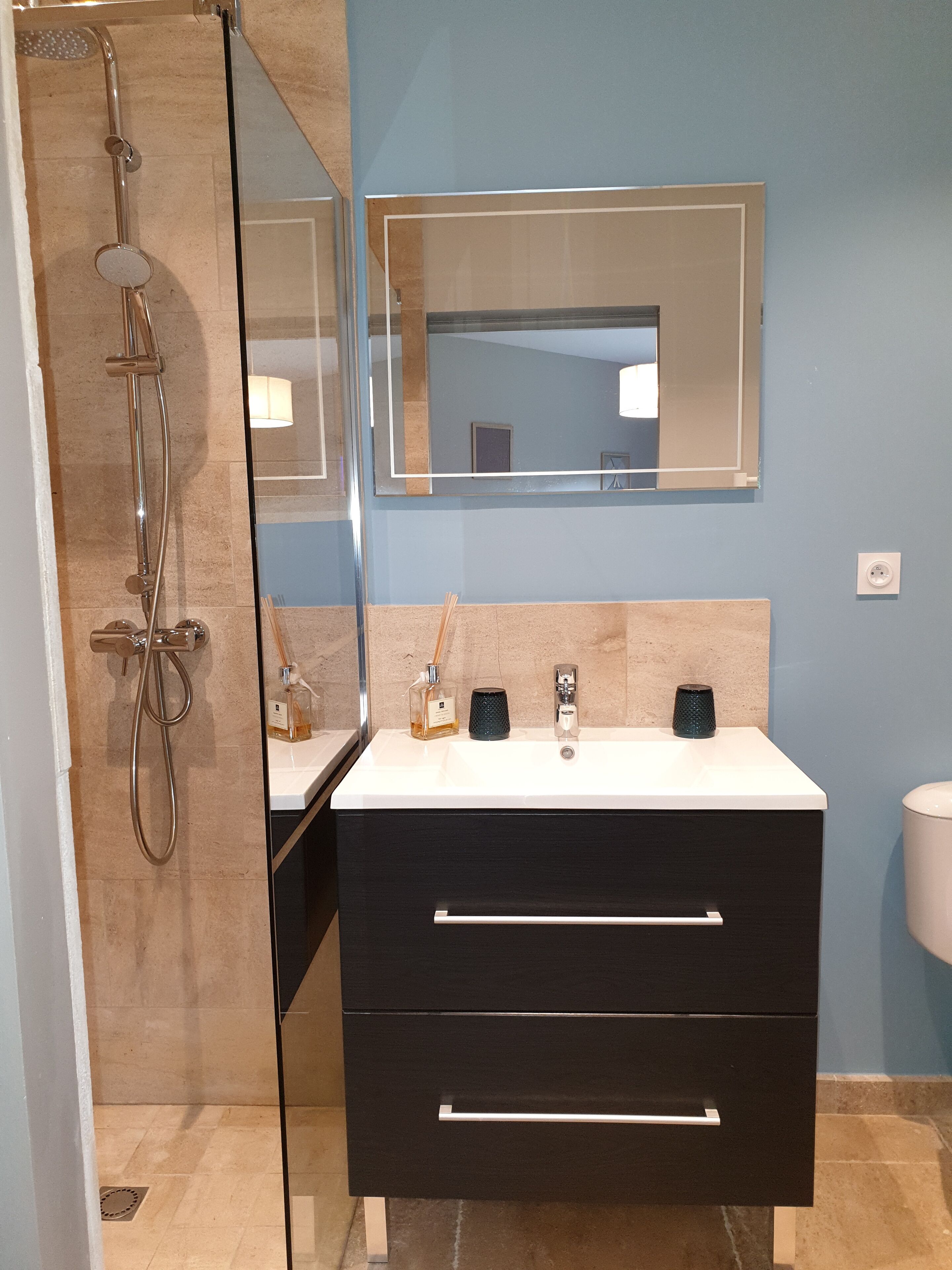 Baño con ducha de vidrio, lavamanos blanco con dos dispensadores de jabón negros, espejo sobre el lavamanos y tocador negro con dos cajones.