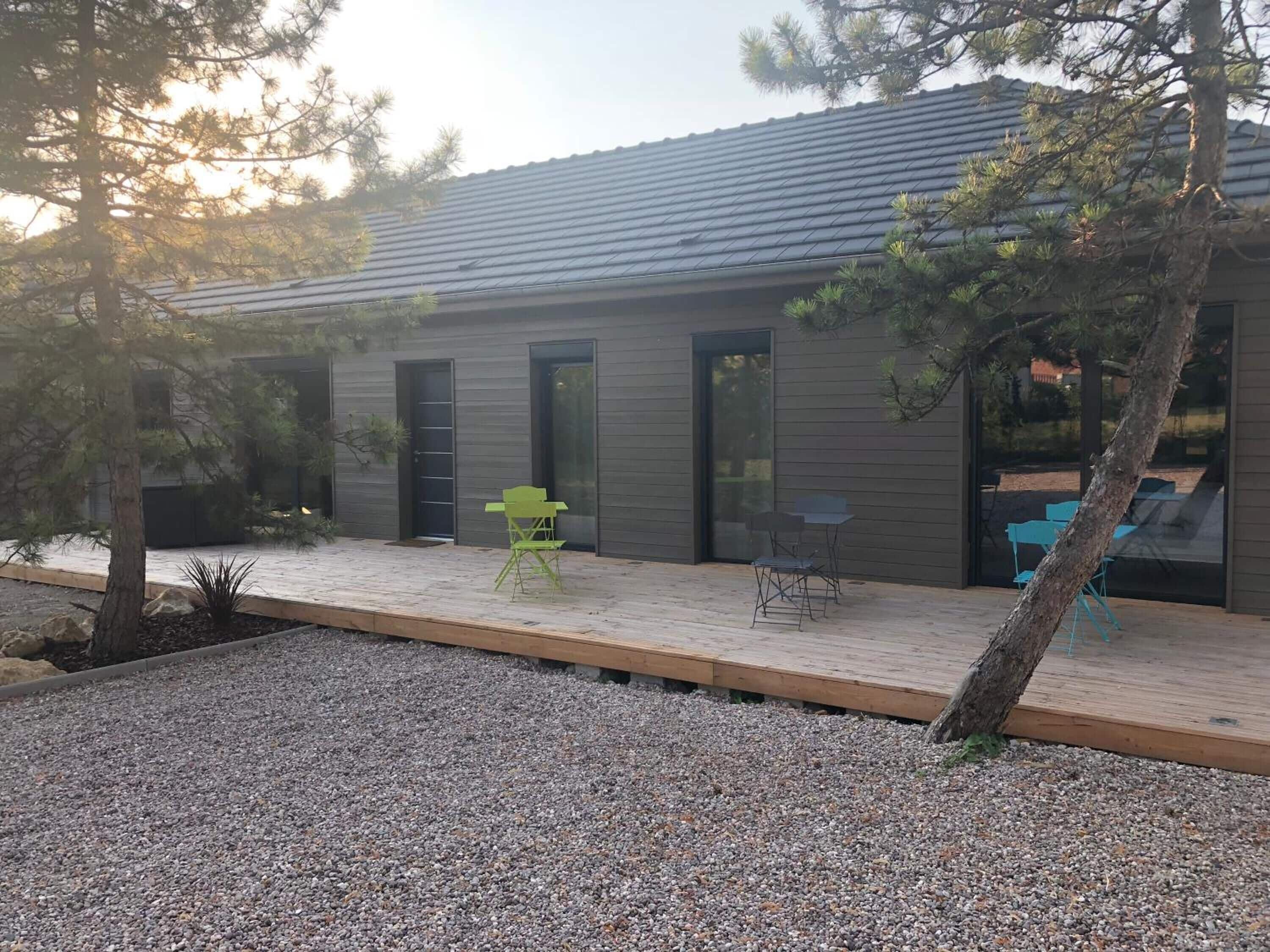 Una casa moderna con una terraza de madera, un pequeño espacio al aire libre con dos sillas y un camino de grava que lleva a la entrada.