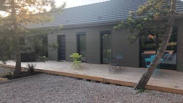 Una casa moderna con una terraza de madera, un pequeño espacio al aire libre con dos sillas y un camino de grava que lleva a la entrada.