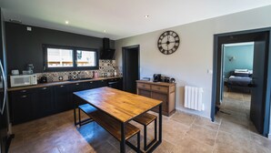 A modern kitchen with dark cabinetry, a wooden table, and a large wall clock. - Cosy Wood House (Laubressel)