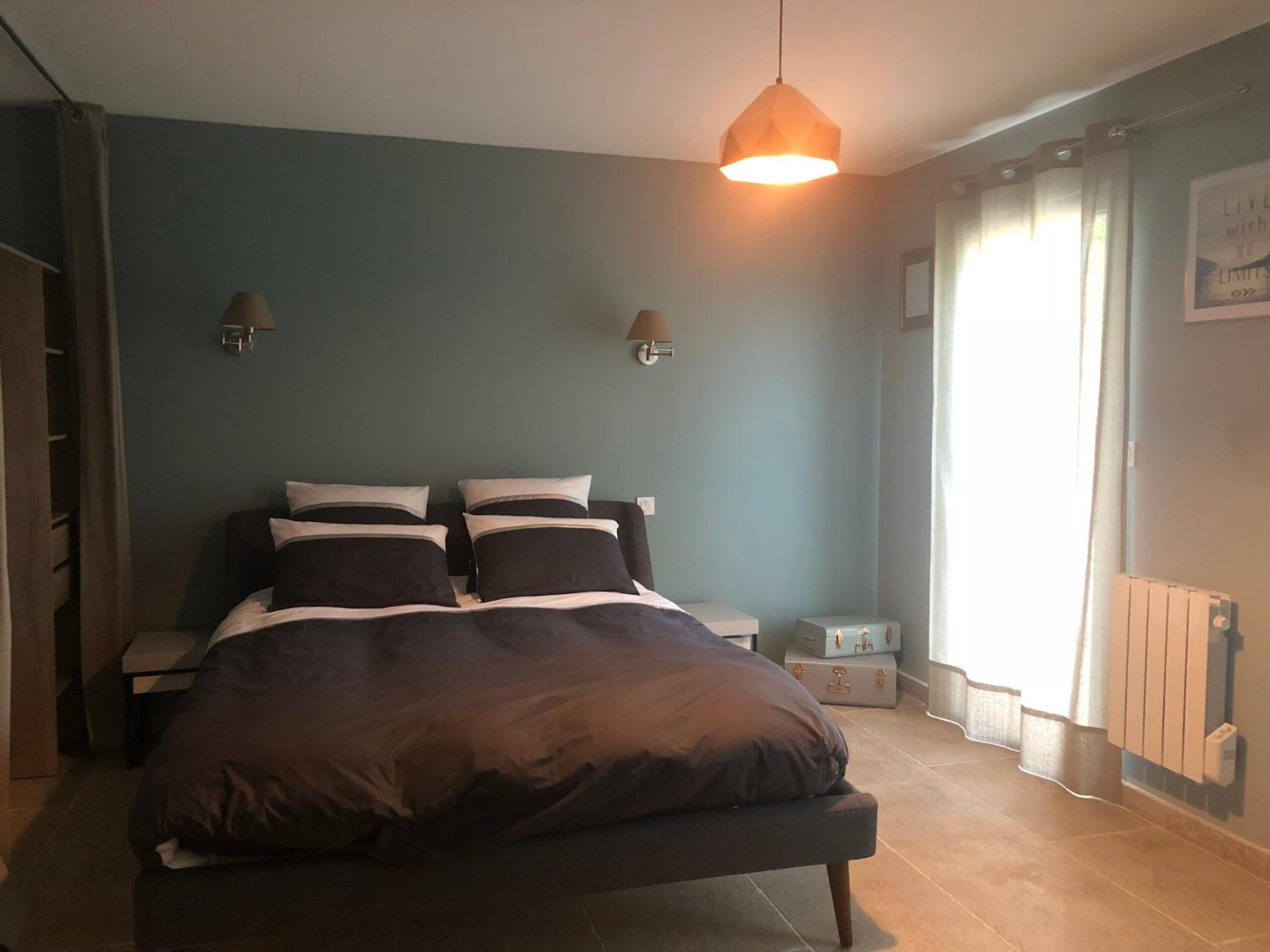 A bedroom with a bed, bedside tables, a nightstand, a window with curtains, and a wall-mounted light fixture.