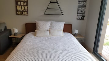 Un lit bien fait, recouvert d'une couette à pois blancs et noirs, avec deux oreillers et une tête de lit en bois. De chaque côté du lit, il y a une table de chevet avec une lampe. Le mur est décoré d'un motif triangulaire et porte une pancarte indiquant les principaux points d'intérêt de la région.