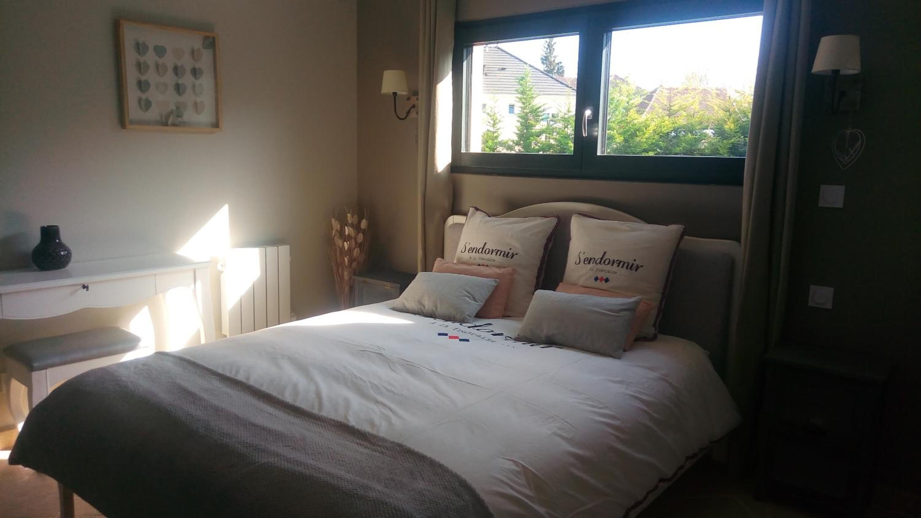 A bedroom with a bed, bedside table, lamp, and a window with a view of trees and buildings.