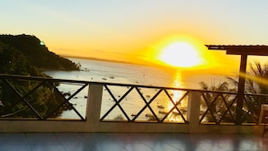 Une vue sur le coucher de soleil depuis un balcon doté d’une balustrade, donnant sur une étendue d’eau où naviguent des bateaux, avec une côte lointaine en arrière-plan.