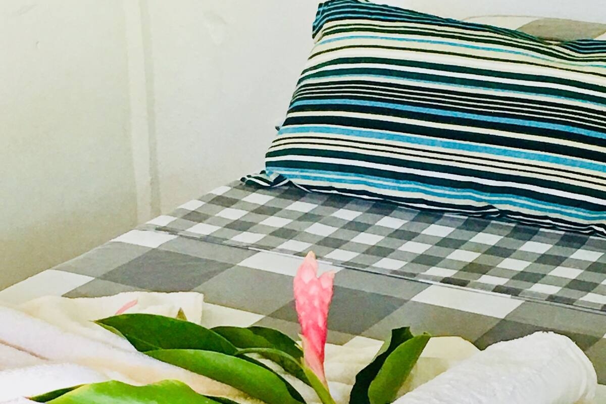 Una cama con colcha de cuadros, almohada a rayas y toallas enrolladas adornadas con hojas verdes y una flor rosa.