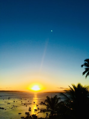 Um pôr do sol sobre a água, com barcos e palmeiras recortadas no céu.