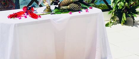 Una mesa adornada con frutas tropicales, un centro de mesa decorativo y vistas al mar con barcos.