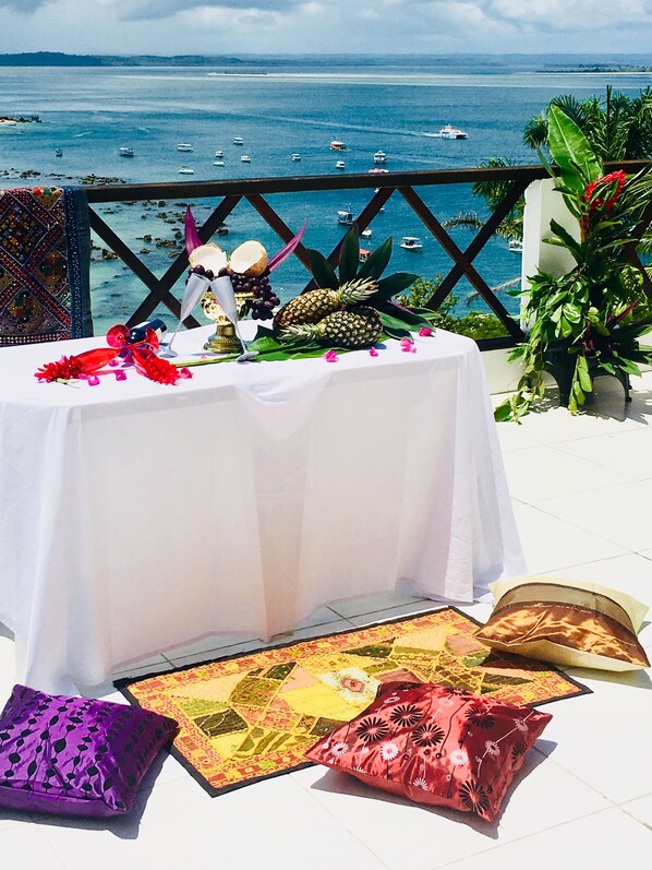 Mesa posta com frutas tropicais, um arranjo decorativo e vista para o mar com barcos.