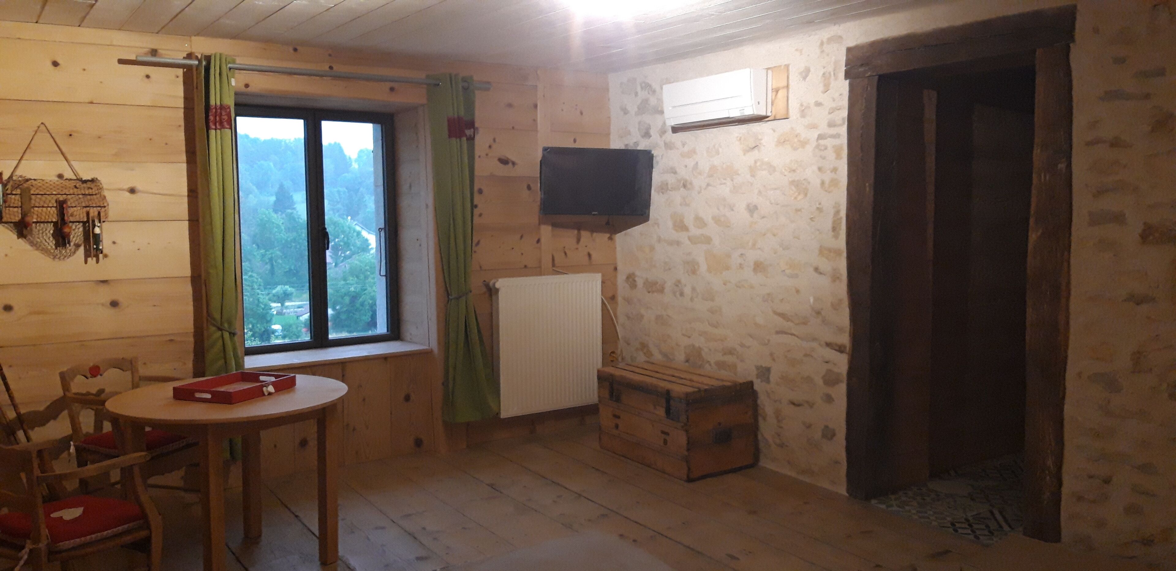 Una habitación con paredes de madera, una ventana con cortinas, una mesa con una caja, un radiador, un televisor y un cofre de madera.