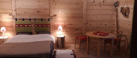 A wooden cabin room with a bed, two chairs, a small table, and a lamp.