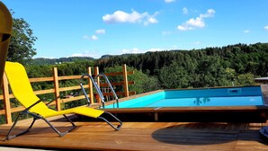 Una terraza de madera con piscina, una silla amarilla y un cerco de madera que da a una ladera boscosa.