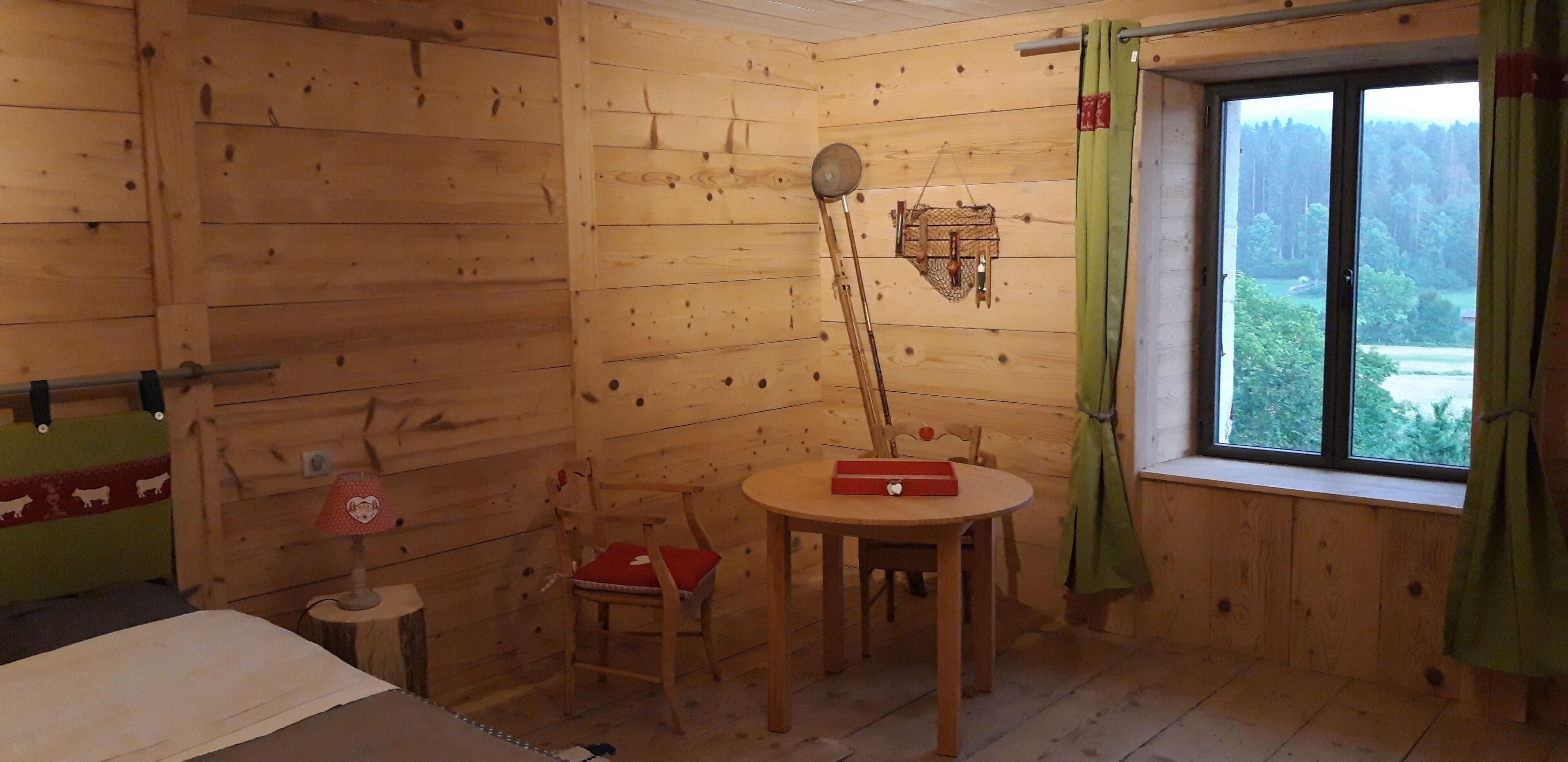 A wooden cabin room with a bed, a small table with a red box, a chair with a red cushion, a wooden wall, and a window with a view of trees.