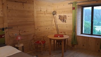 A wooden cabin room with a bed, a small table with a red box, a chair with a red cushion, a wooden wall, and a window with a view of trees.