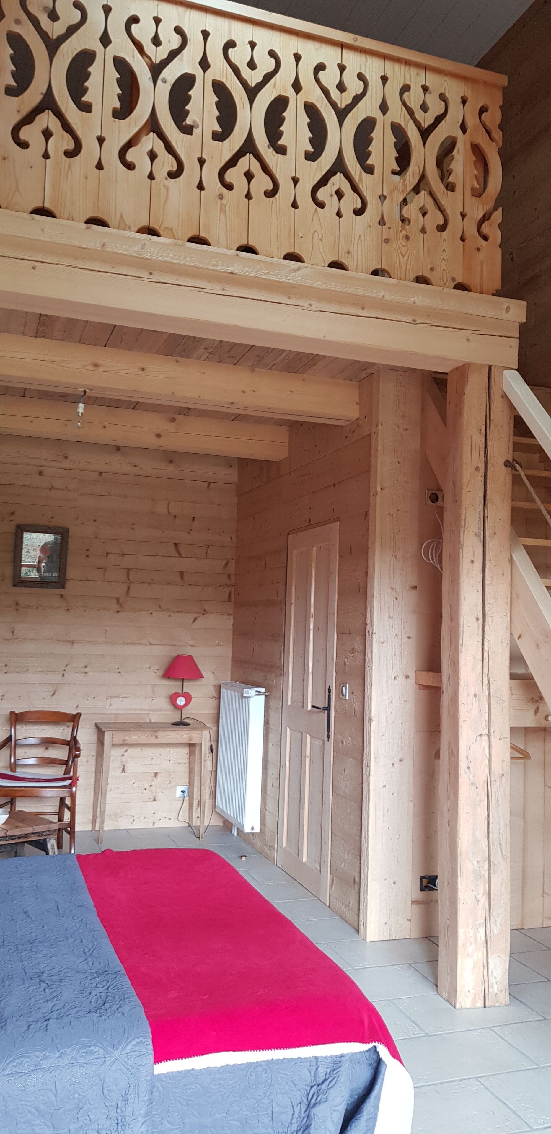 A wooden cabin interior with a bed, a chair, a table with a red lamp, a door, and a staircase.