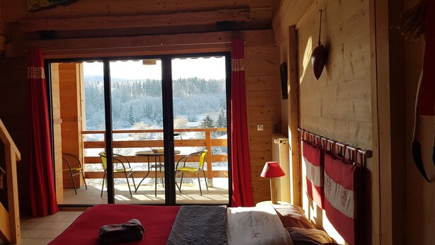 Habitación de cabaña de madera con cama, un tapete rojo, una mesa con sillas y vista a un bosque nevado a través de una puerta corrediza.