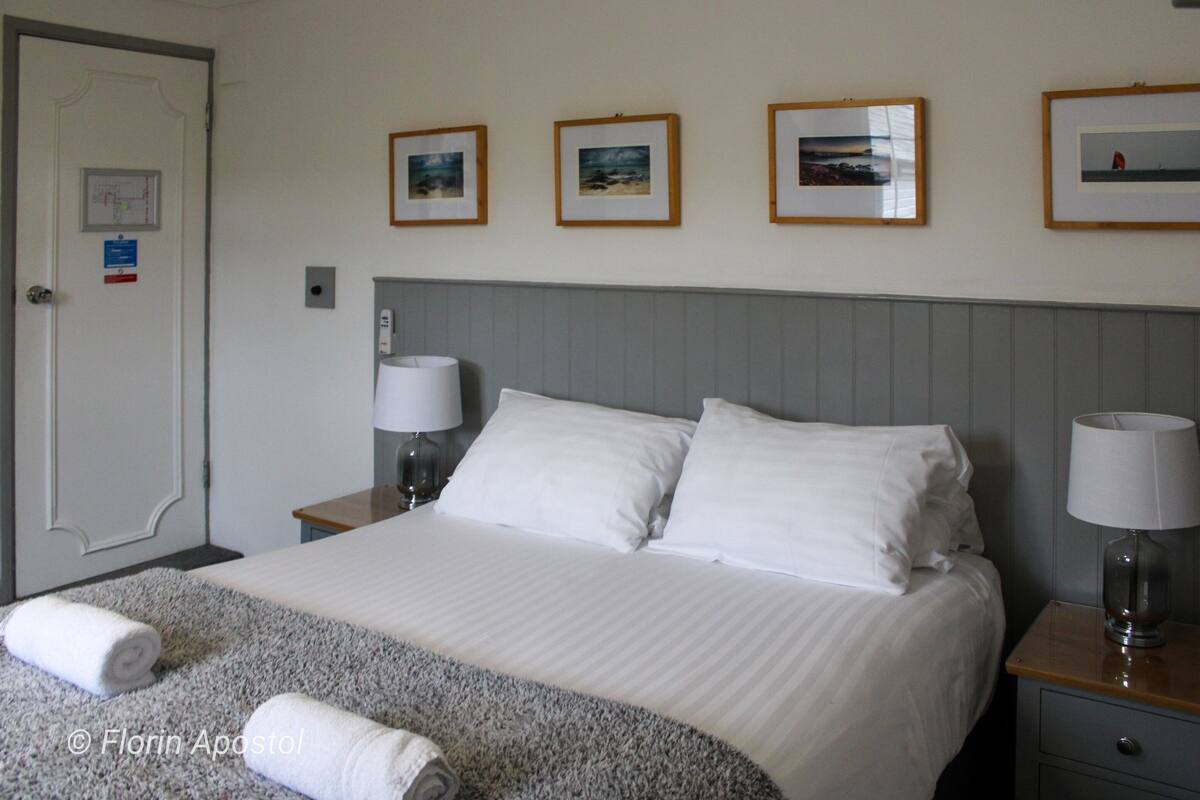 A hotel room with a neatly made bed, two bedside tables with lamps, a door with a map and sign, and three framed pictures on the wall.