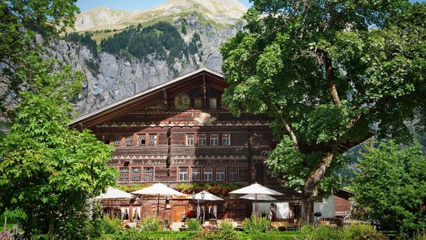A traditional wooden building with a sloped roof, surrounded by lush greenery and a mountain backdrop. - Landgasthof Ruedihus (Kandersteg)