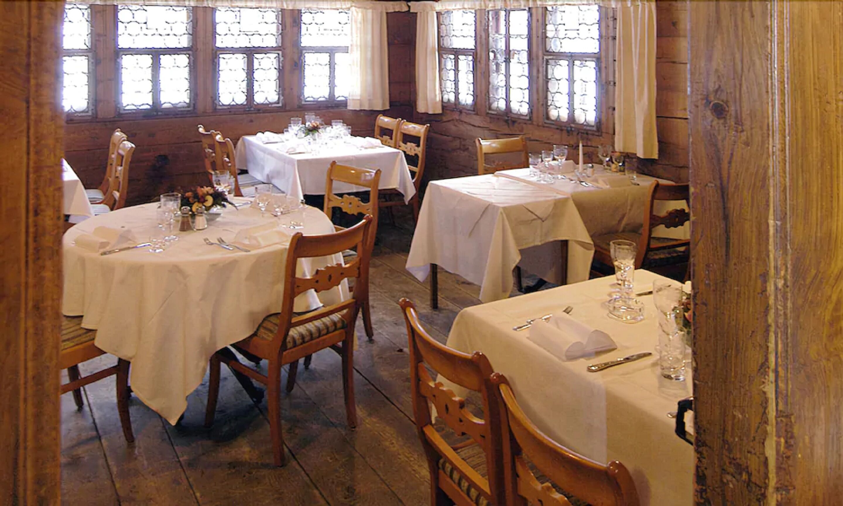 A dining area with round tables covered in white tablecloths, wooden chairs, and large windows with decorative glass panes.