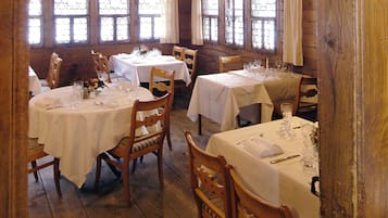 A dining area with round tables covered in white tablecloths, wooden chairs, and large windows with decorative glass panes.
