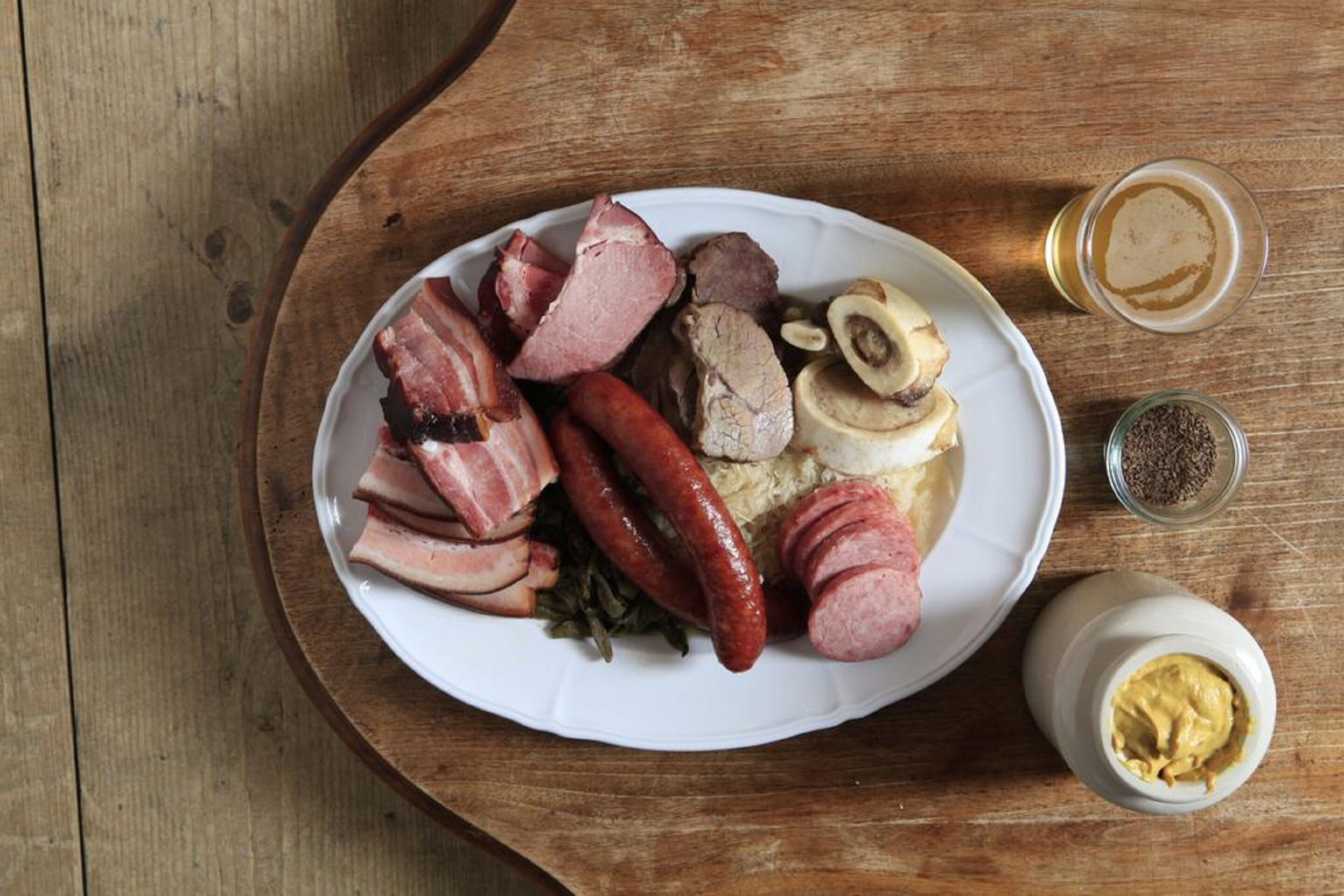 Prato de comida com salsichas, presunto, cogumelos e um pequeno recipiente com mostarda sobre uma mesa de madeira.