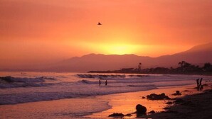 Escena de playa al atardecer con olas, un ave volando y siluetas de personas.
