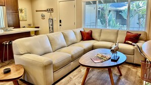Una sala de estar con un sofá seccional color beige, una mesa redonda y una cocina con armarios de madera y un refrigerador blanco.