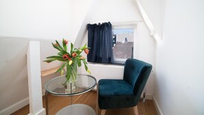 A small, bright room with a teal chair, a glass table with a vase of flowers, and a window with dark curtains. - Dase Bed & Breakfast (Amsterdam)