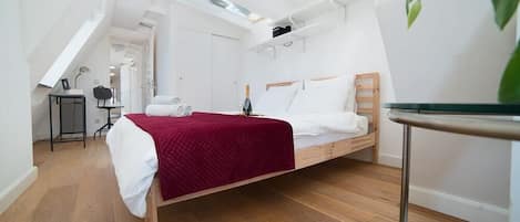 Un dormitorio con cama de madera, ropa de cama blanca, una manta roja, una mesita, una silla y una planta.