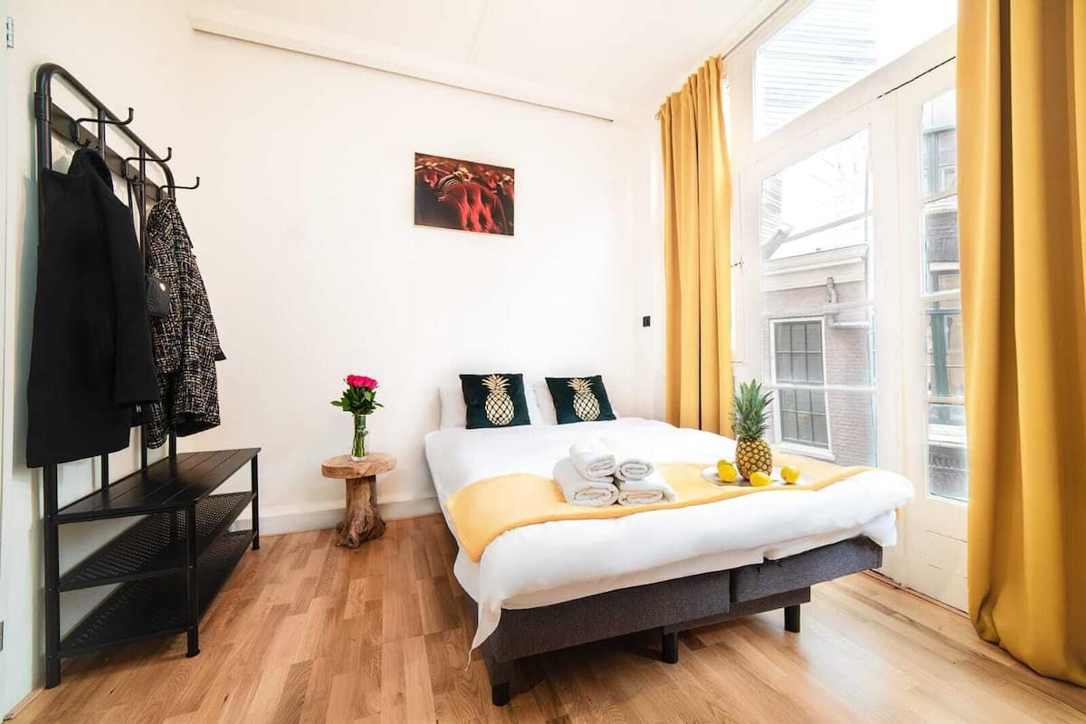 A hotel room with a bed, a small side table with a vase of flowers, a coat rack, and a window with curtains.