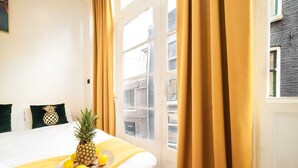 A hotel room with a bed, yellow curtains, a pineapple on the bed, and a view of buildings outside. - Amazing B&B (Amsterdam)