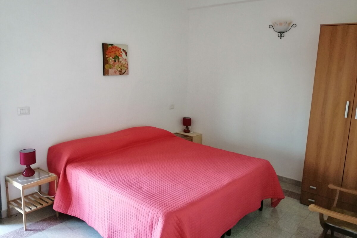 Quarto com uma cama de solteiro, um guarda-roupa de madeira, uma pequena mesa de cabeceira com luminária e um aplique de parede.