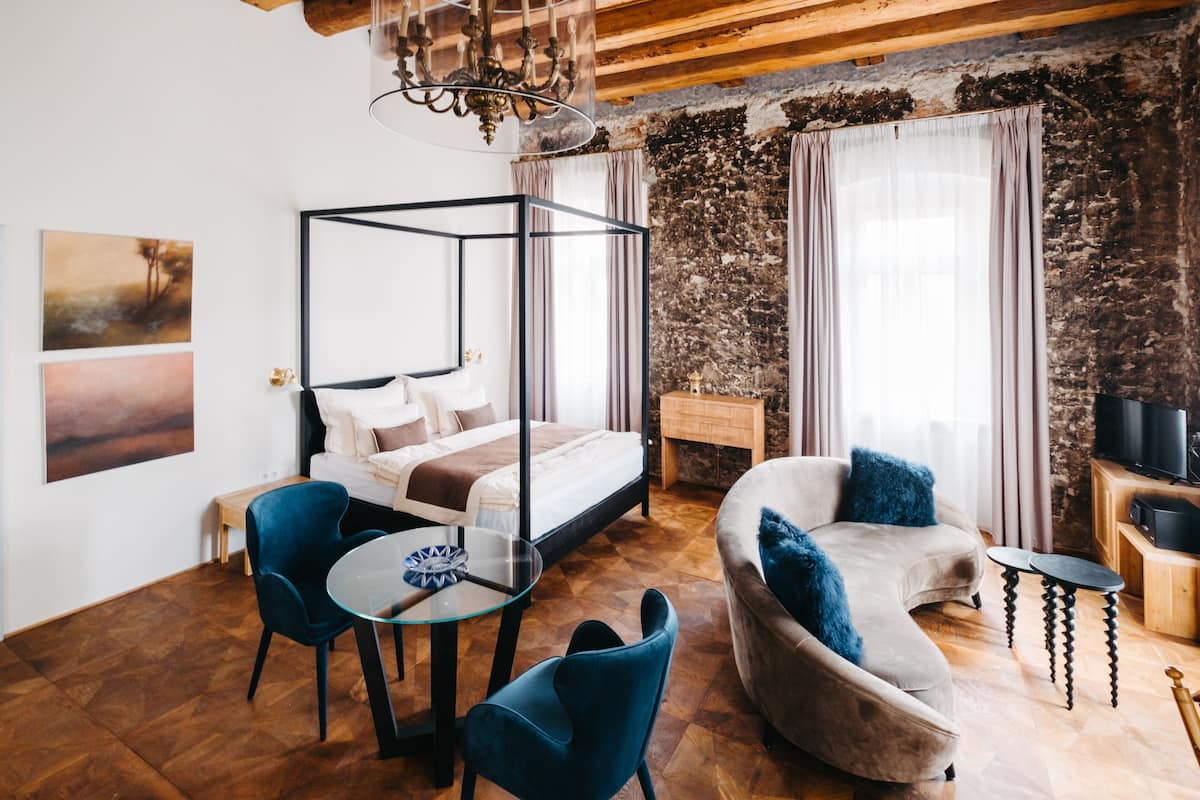 A modern bedroom with a four-poster bed, a round glass table, two blue chairs, a curved sofa with blue cushions, and a stone wall.