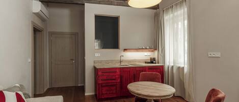 Un espacio habitable compacto con kitchenette, un comedor con mesa redonda, una silla roja y un sofá con cojín rojo.