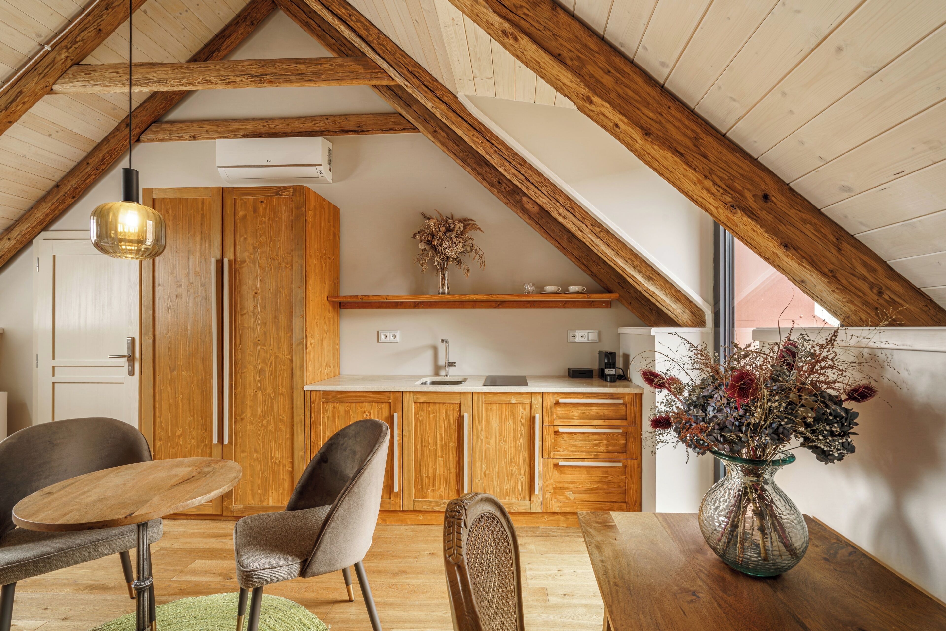 A loft-style room with a wooden ceiling, a dining table, two chairs, a vase with dried flowers, and a kitchen area with wooden cabinets and a countertop.
