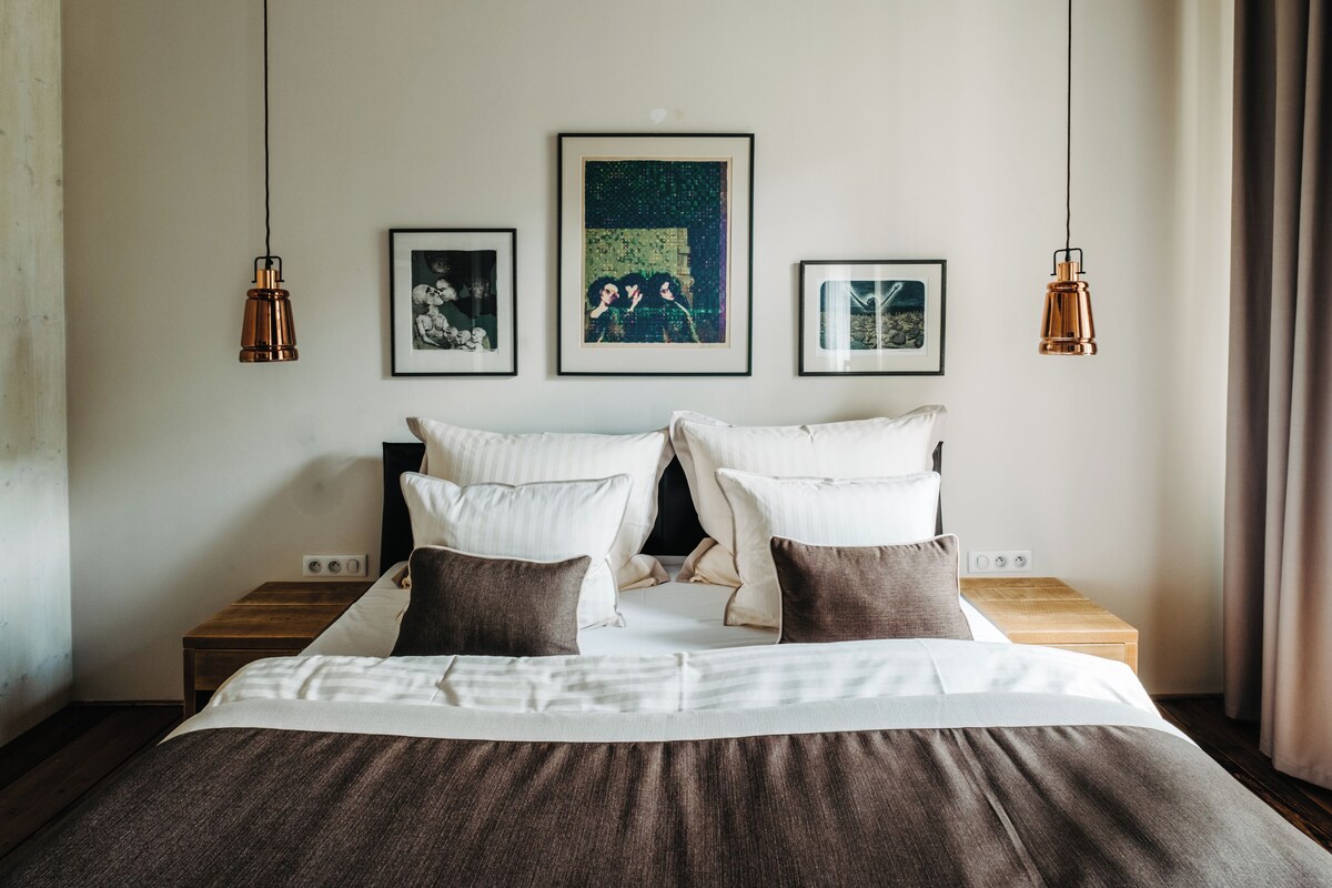 Una cama bien hecha con dos almohadas, una mesita de noche con una lámpara y tres cuadros enmarcados en la pared.