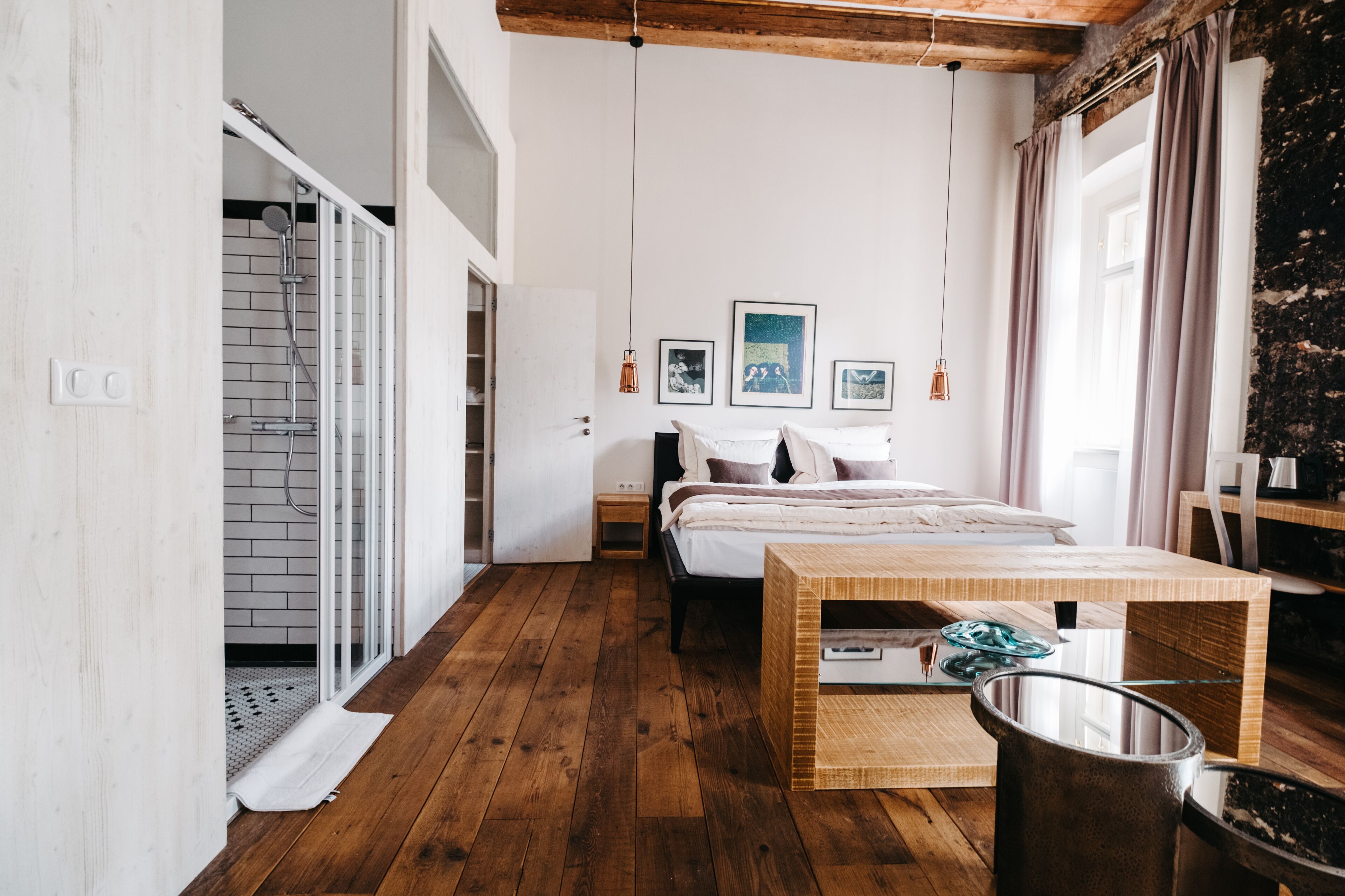 A bedroom with a wooden floor, a bed, a bedside table, a nightstand, a window with curtains, and a shower area.