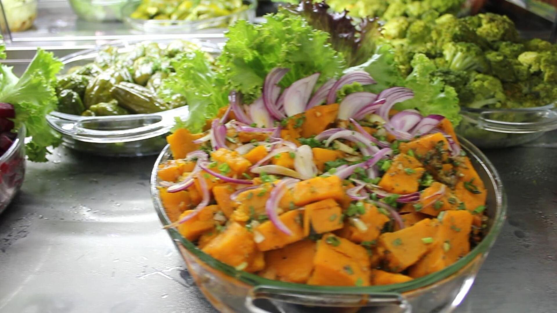 Un saladier en verre rempli d’une salade composée de patates douces coupées en dés, d’oignons rouges et d’herbes aromatiques, entouré d’autres saladiers.