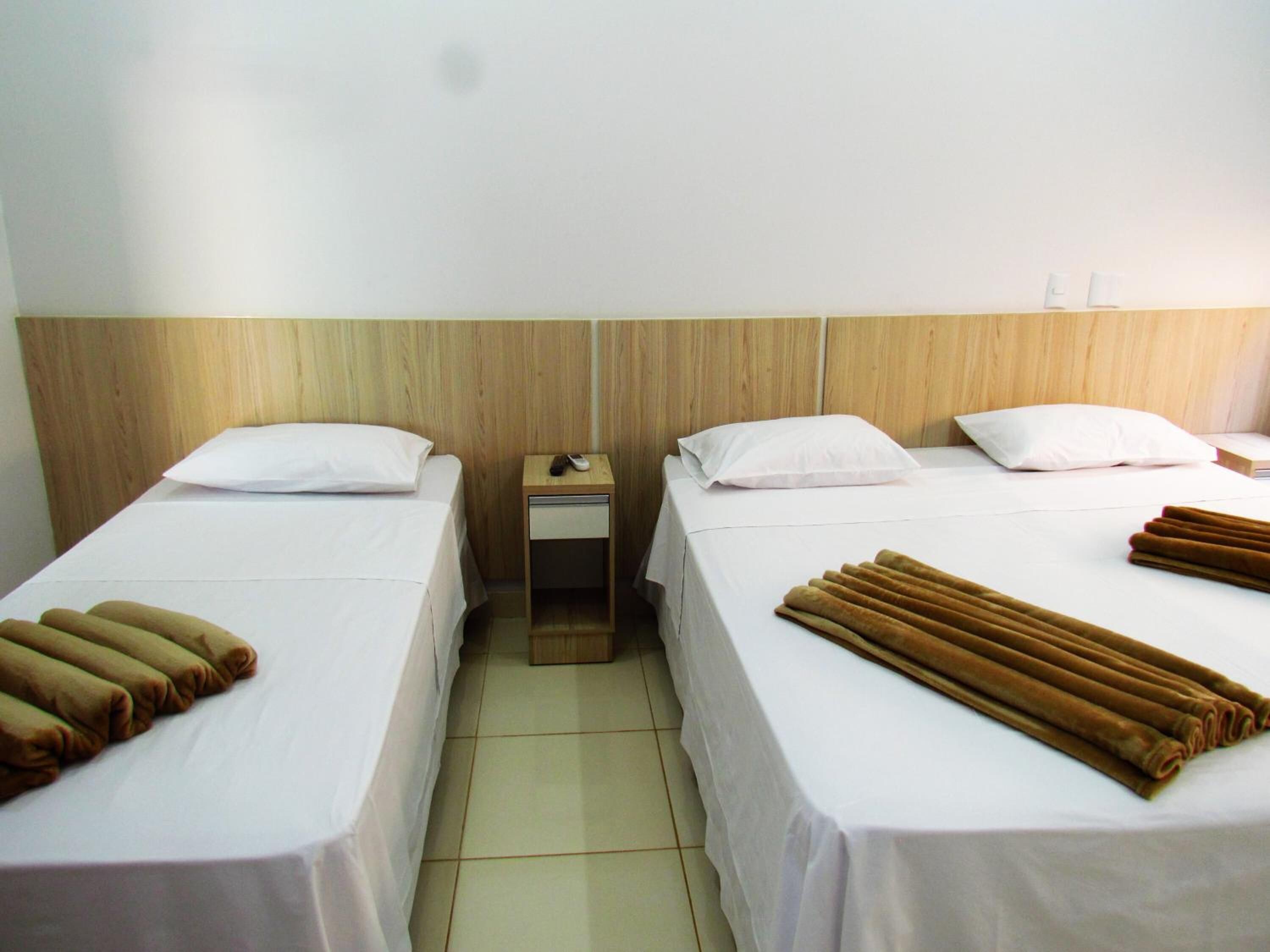 A hotel room with two single beds, each with a white pillow and a brown blanket. There is a wooden headboard, a nightstand with a phone, and a small table with a book.