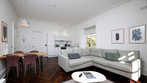 Ein modernes Wohnzimmer mit einem grauen Ecksofa, einem Esstisch mit Stühlen, einer Küche im Hintergrund und Bildern an den Wänden.